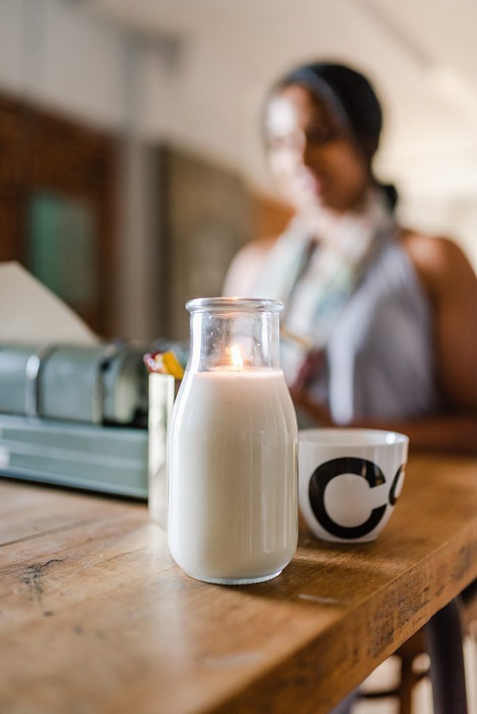 glass bottle of milk and a small white cup on a wooden table. a woman using the typewriter can be seen in the background but they're out of focus