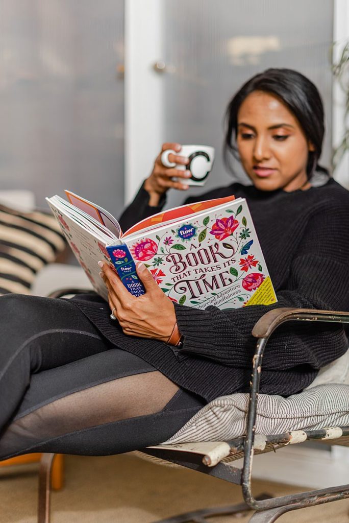 woman in black sweater is lounging on an arm chair while reading a book called "a book that takes it's time" snd holding a white cup in one hand
