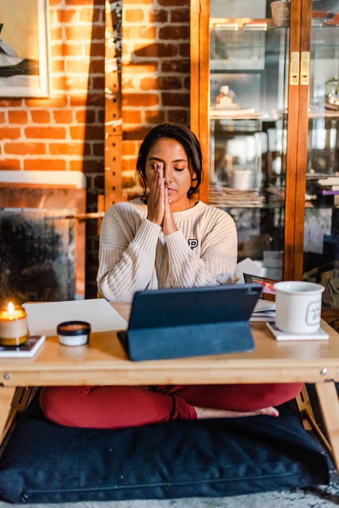 woman in white sweater is sitting cross legged by a small table. she has her laptop in front of her plus a big mug and a lit candle on either sides of it. she has her hands clasped together in front of her face while her eyes are closed.