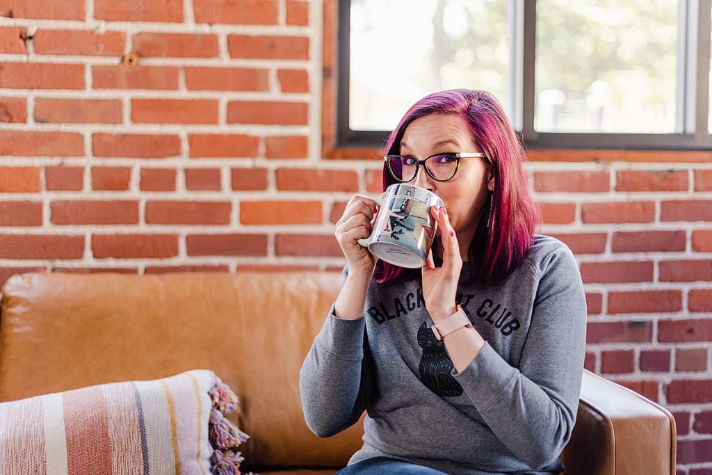 woman with pink hair wearing a gray sweater is drinking from a white mug while sitting on a brown leather couch