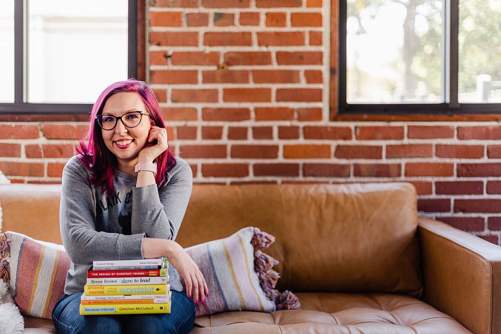 woman with pink hair and glasses is sitting on a brown leather sofa. she has a stack or books in her lap and she is leaning on them.