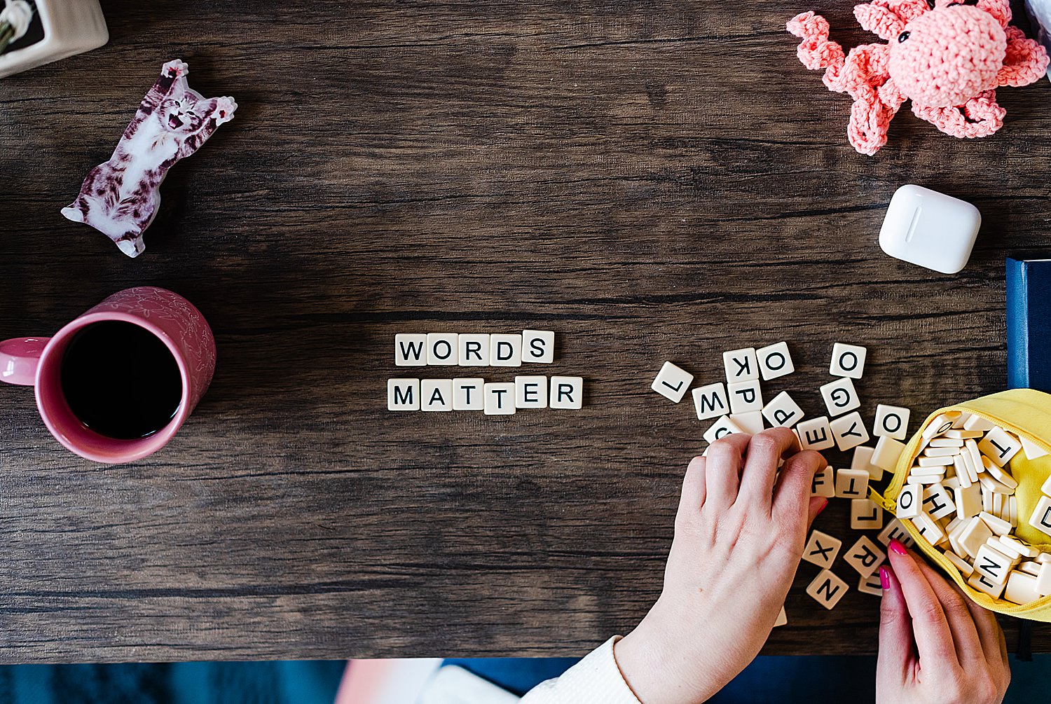 flatlay image of a wooden tabletop with letter tiles that spells out "WORDS MATTER" in the middle. there's a couple of scattered tiles and some in a yellow pouch on the lower right hand side with two hands picking up some tiles, above that there's an airpods case and a knitted coral-colored octopus and on the left side of the photo is a pink mug with black coffee