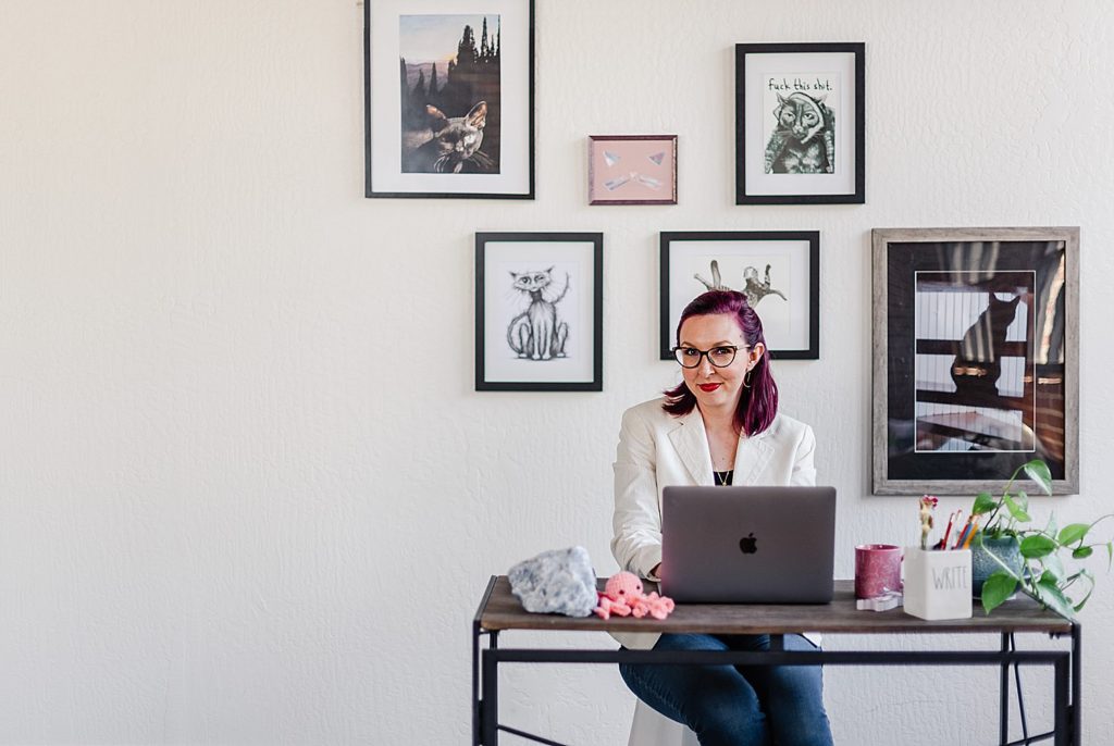 woman in white suit jacket sitting in front of her desk with her laptop opened in front of her. the wall behind her has some framed cat photos and art of different sizes