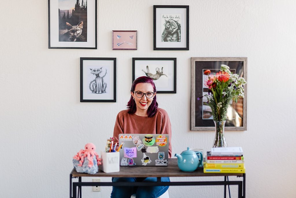 a woman wearing a sweater is sitting in front of her desk with her laptop opened in front of her. the wall behind her has some framed cat photos and art of different sizes