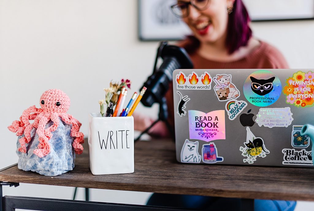 a laptop with lost of different stickers on it, a white pencil holder that says "WRITE", and a knitted octopus on top of a rock are all on top of a wooden desk. on the background, a woman can be seen sitting in front of the desk while speaking on a mic