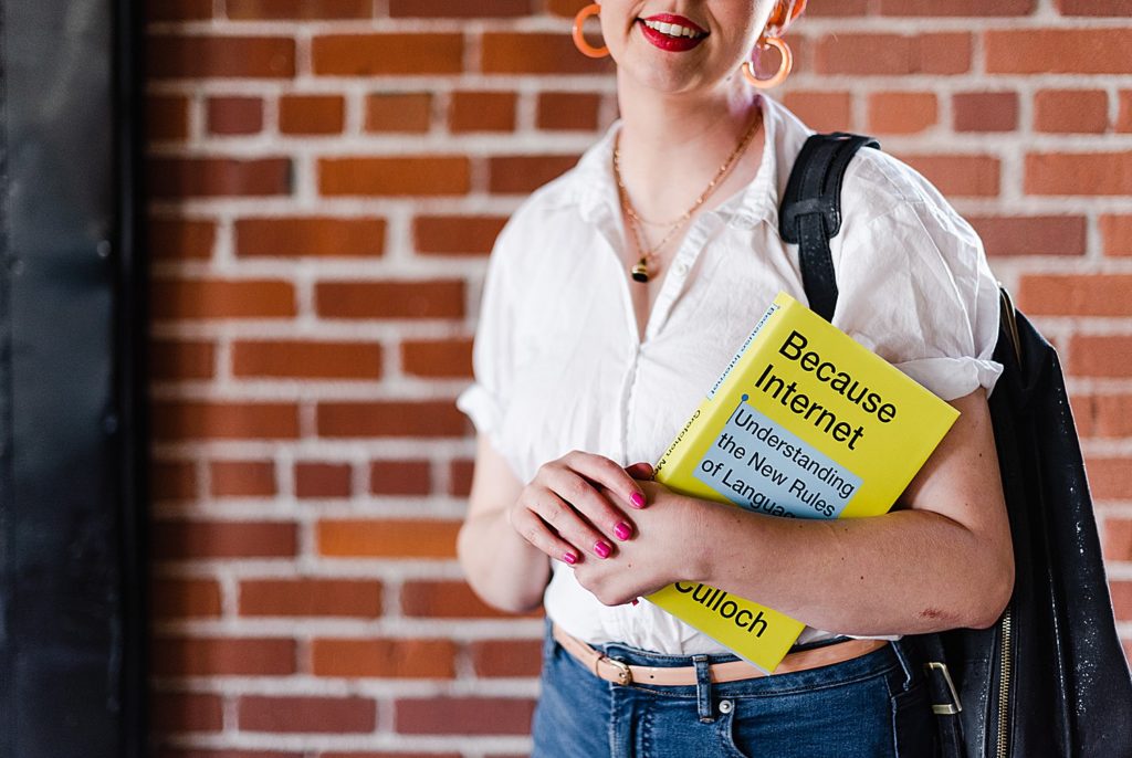 woman in a white blouse with a black backpack hanging on one shoulder is holding a yellow book called "Because Internet"