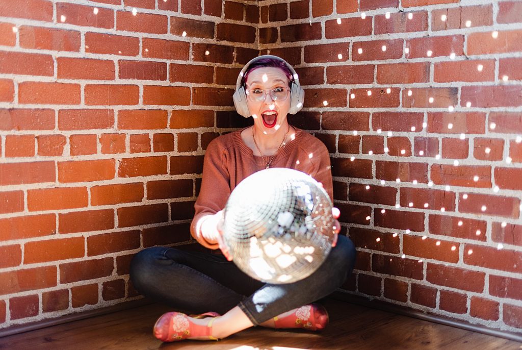 woman in brown sweater sitting indian style wearing headphones while holding a disco ball. the background is a brick wall and the woman looks like she's screaming in delight
