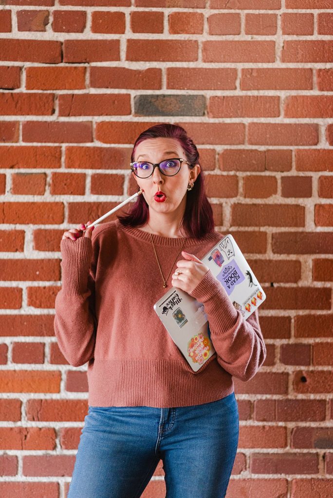 a woman in brown sweater is standing in front of a brick wall. she's holding her laptop with one hand and a pencil pointing to her fake shocked face with the other