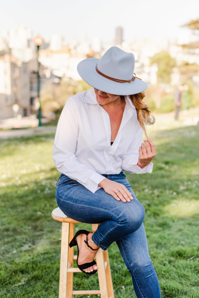 woman in white long-sleeved shirt and jeans sitting on a wooden stool wearing a blue cowboy with her face partially hidden by the hat