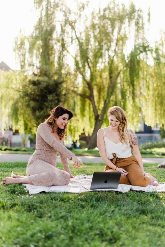 two women sitting on a picnic blanket in a park, they're looking at the laptop in front of them. the woman on the left is pointing to the laptop screen