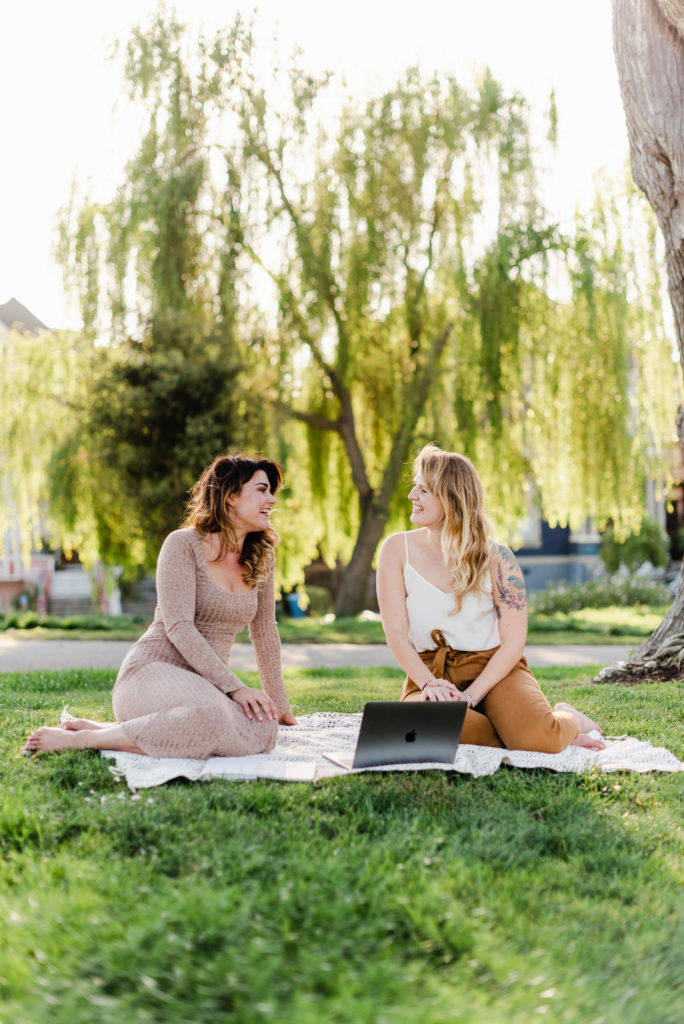 two women sitting on a picnic blanket in a park looking at each other smiling. there's an open laptop in front of them