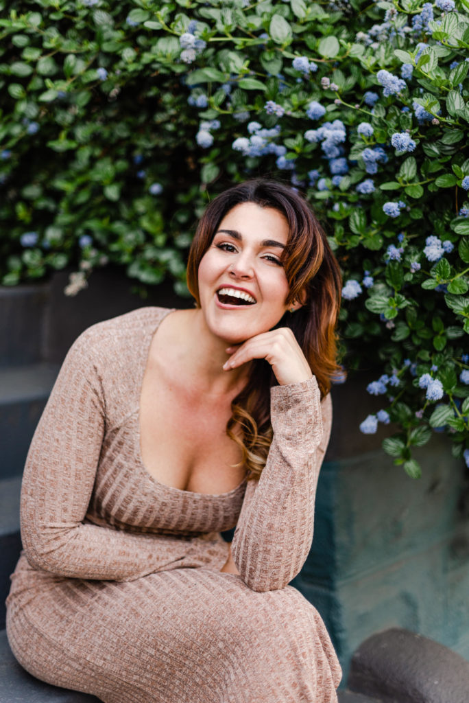 woman sitting in front of a flower bush. she is smiling while leaning her face on her hand