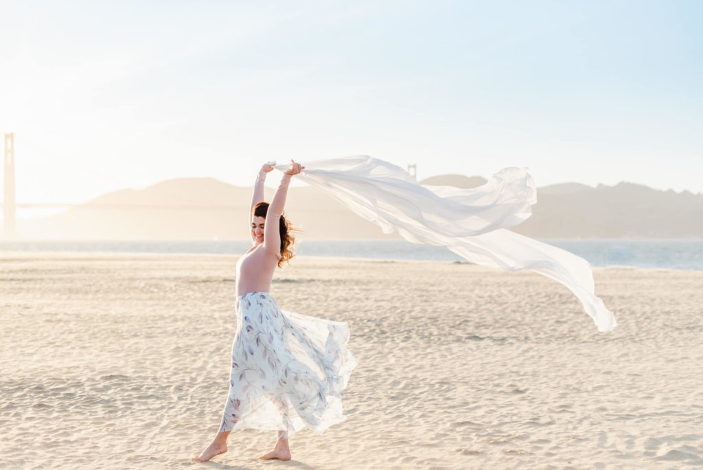woman in a flowy white skirt is holding a white blanket on her raised arms. her skirt and the blanket are being blown by the wind
