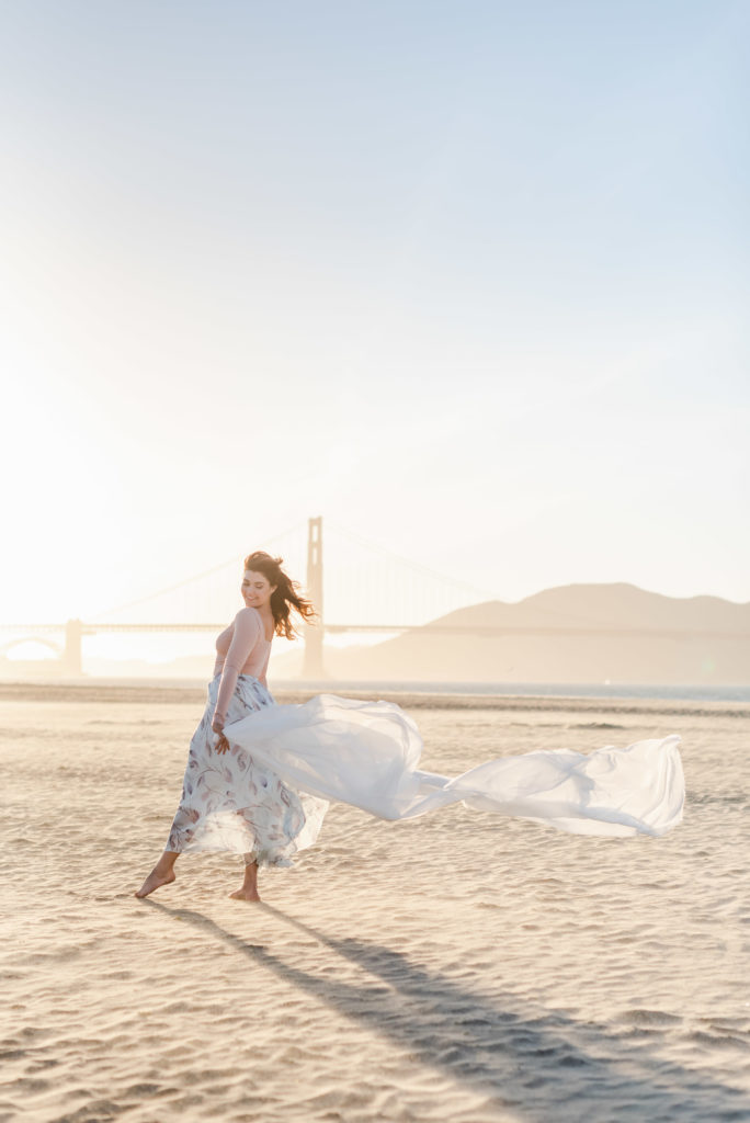 woman in a flowy white skirt is holding a white blanket behind her and letting it be blown by the wind along with her skirt