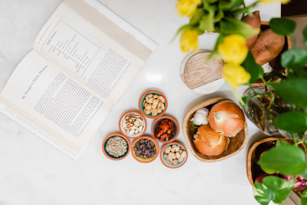 flatlay of 6 different spices on brightly colored saucers arranged in a pyramid, two white onions and a garlic bulb on a wooden, square saucer, a wooden coaster above it and an open book to the left of everything. the book is open on a chapter called "Dr. Greger's Daily Dozen".
