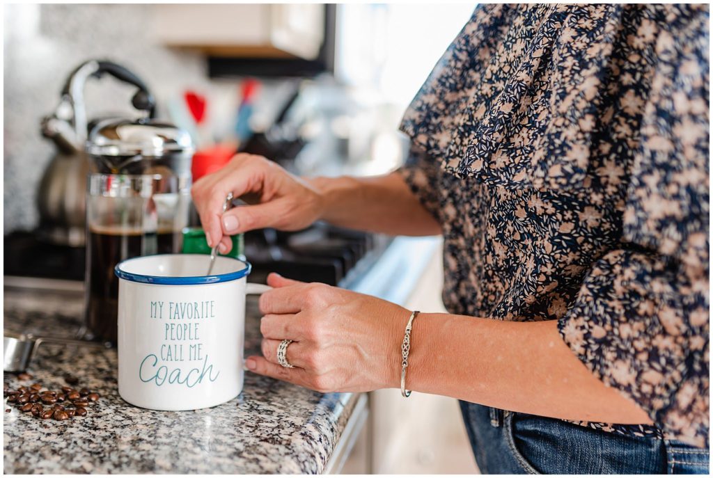 a white mug with the words "my favorite people call me coach" on it on a countertop; a woman is holding the mug with one hand and stirs it with the other