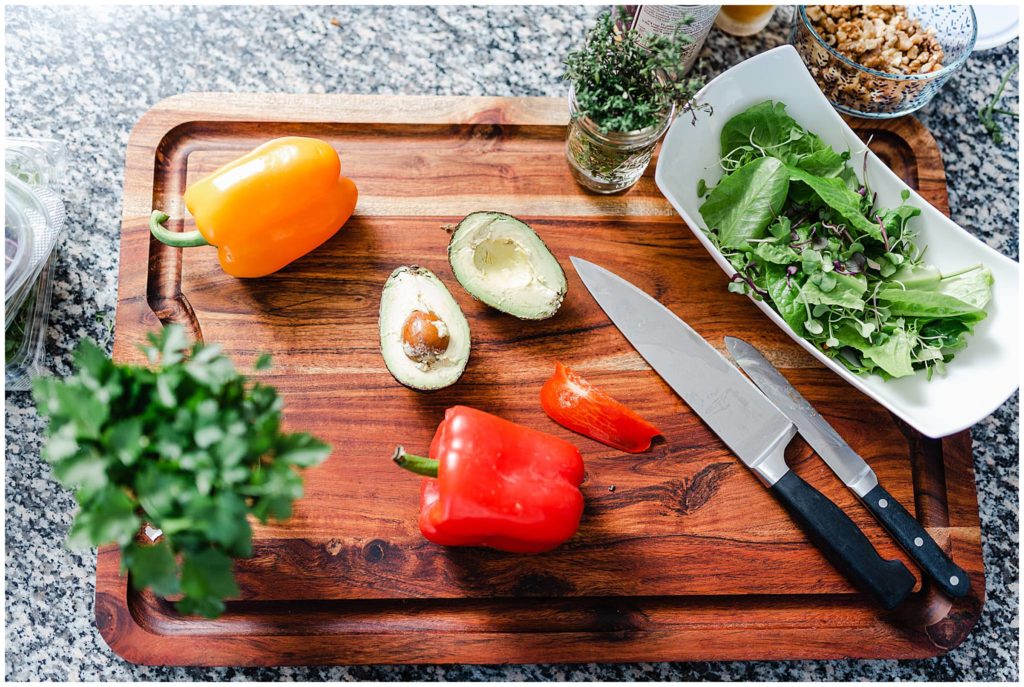 flatlay image of bell peppers, avocado, leafy greens, a cooking knife and abutter knife on top of a wooden cutting board