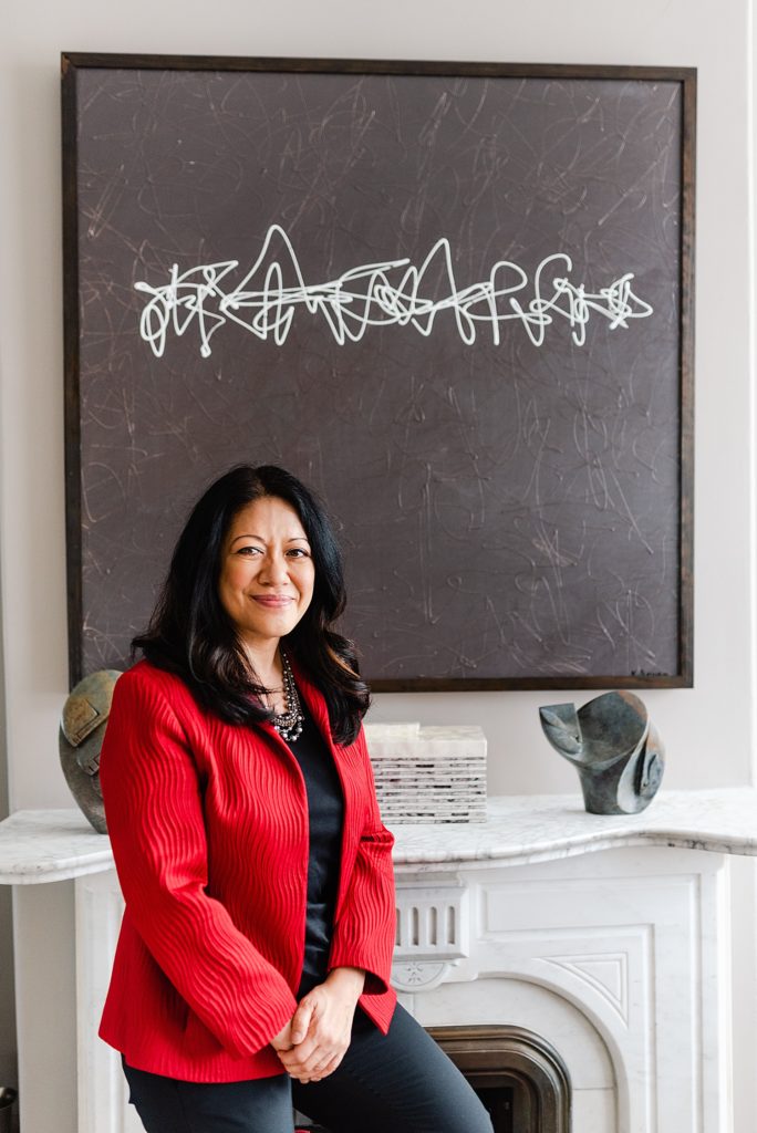 woman in red blazer is standing by a fireplace with a black and white abstract artwork above the mantel