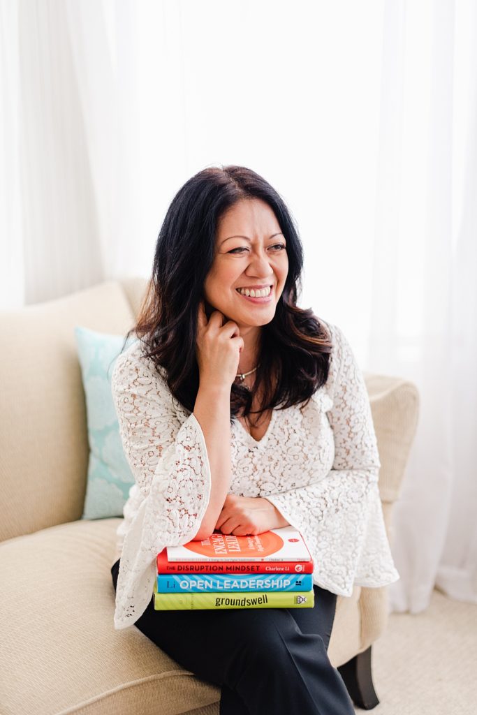 woman in white lace blouse is sitting on a couch with a stack of book on her lap. she is smiling but is looking to the side and not at the camera