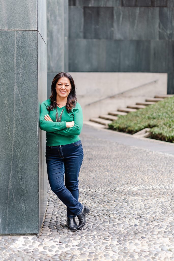 woman in green long-sleeved shirt and jeans has her arms crossed and is leaning on a stone column