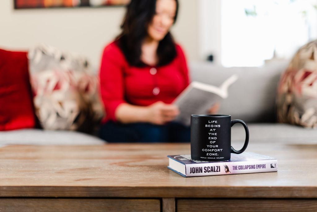 black mug on top of a book on a wooden tabletop. the mug has writing that says "Life begins at the end of your comfort zone. -Neale Donald Walsch"; the book is called "The Collapsing Empire" by John Scalzi