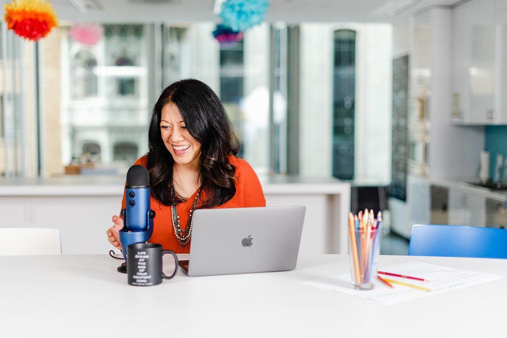 smiling woman with a blue mic, a macbook, and a black mug on the table in front of her. there's also a glass full of colored pencils and some papers to the woman's left side