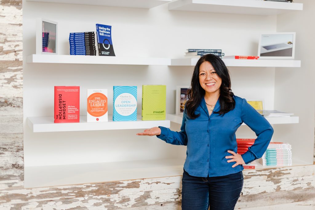woman in blue blouse showing off some books on a shelf beside her. her hand is gesturing to the books beside her