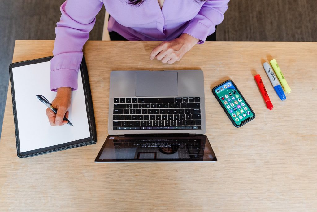 flatlay image of a wooden desk with a laptop, an iphone, three highlighters in different colors, and a pad of paper with a woman's hand holding a pen on top of it.