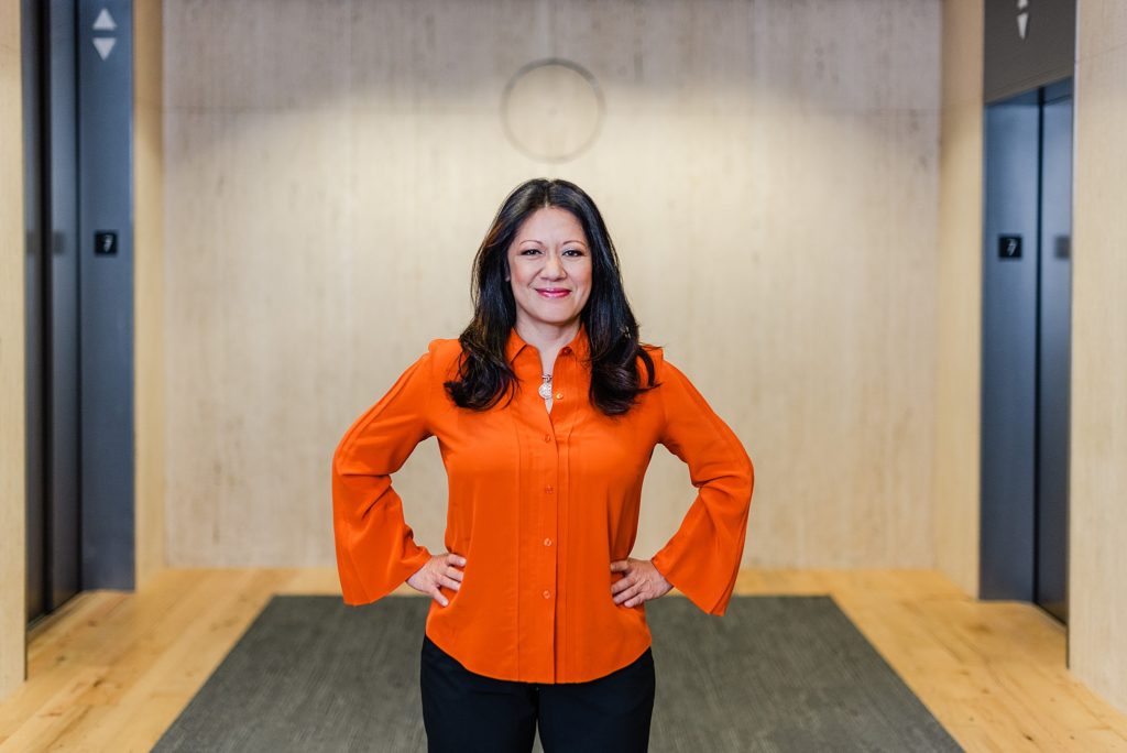 woman in orange long-sleeved blouse has both her hands on her hips and is smiling to the camera