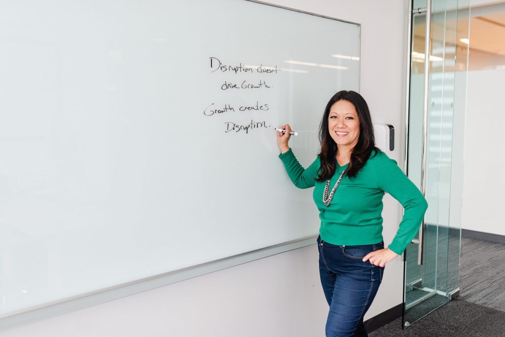 woman in green is holding a marker to the white whiteboard and is holding her hip with her other hand. the words "disruption doesn't drive growth. growth creates disruption." is written on the whiteboard.