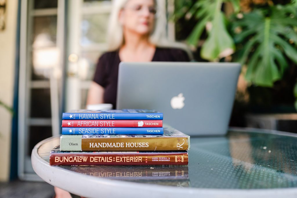 a stack of books about house decoration on top of a glass round table. there is a woman working on her laptop in the background