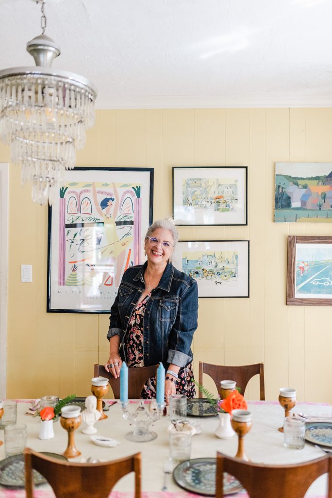 woman standing in a decorated dining area. the wall behind her has a bunch of colorful artworks and paintings