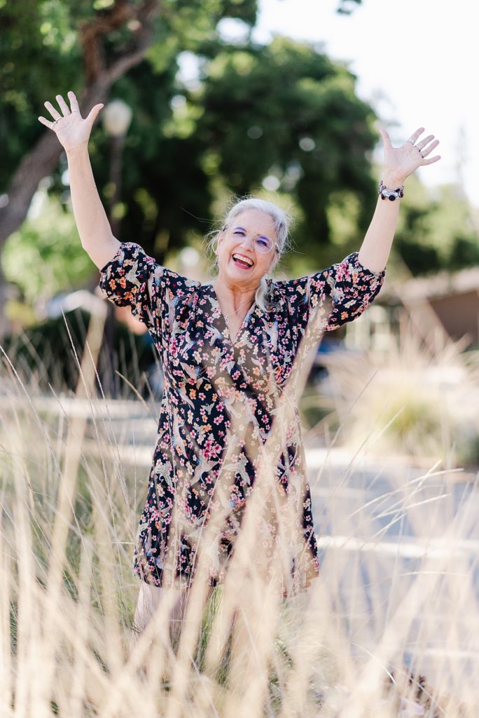 laughing woman in a floral dress raising her hands up. there's grass around her, in the background and foreground of the image
