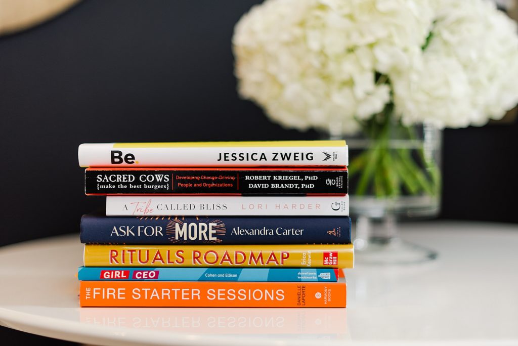 stack of books on a white table. Be by Jessica Zweig;Sacred Cows (make the best burgers) by Robert Kriegel, PhD and David Brandt, PhD; A Tribe Called Bliss by Lori Harder; Ask For More by Alexandra Carter; Rituals Roadmap by Erica Keswin; Girl CEO by Cohen and Ellison; and The Fire Starter by Danielle Laporte
