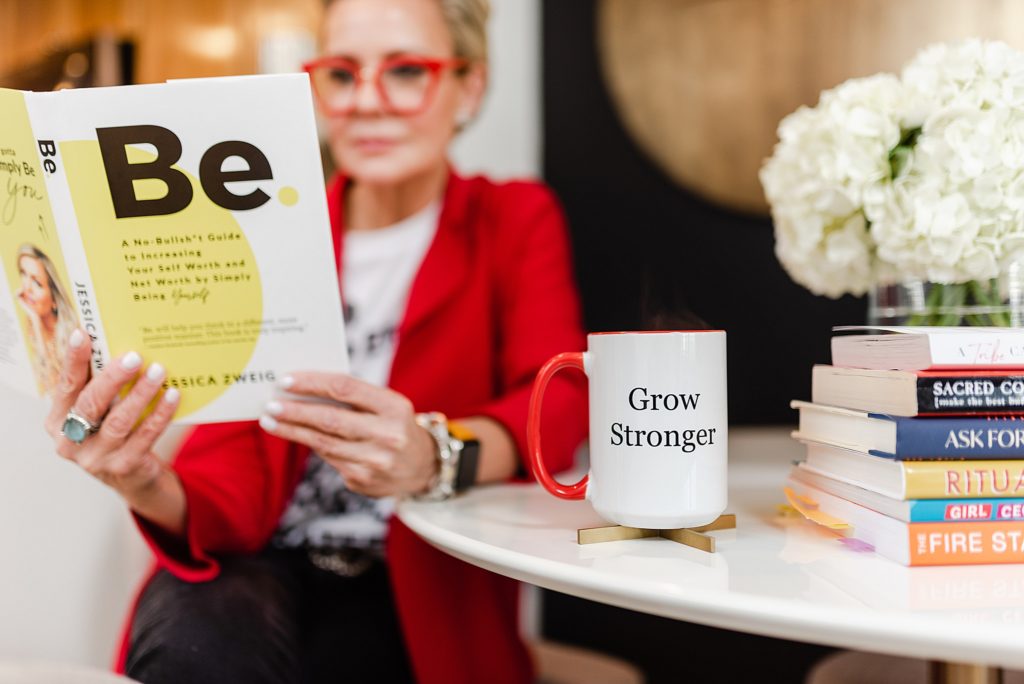 White mug with red handle has the words "Grow Stronger" on it. Woman in red blazer sitting beside the table is reading the book "Be" by Jessica Zweig.