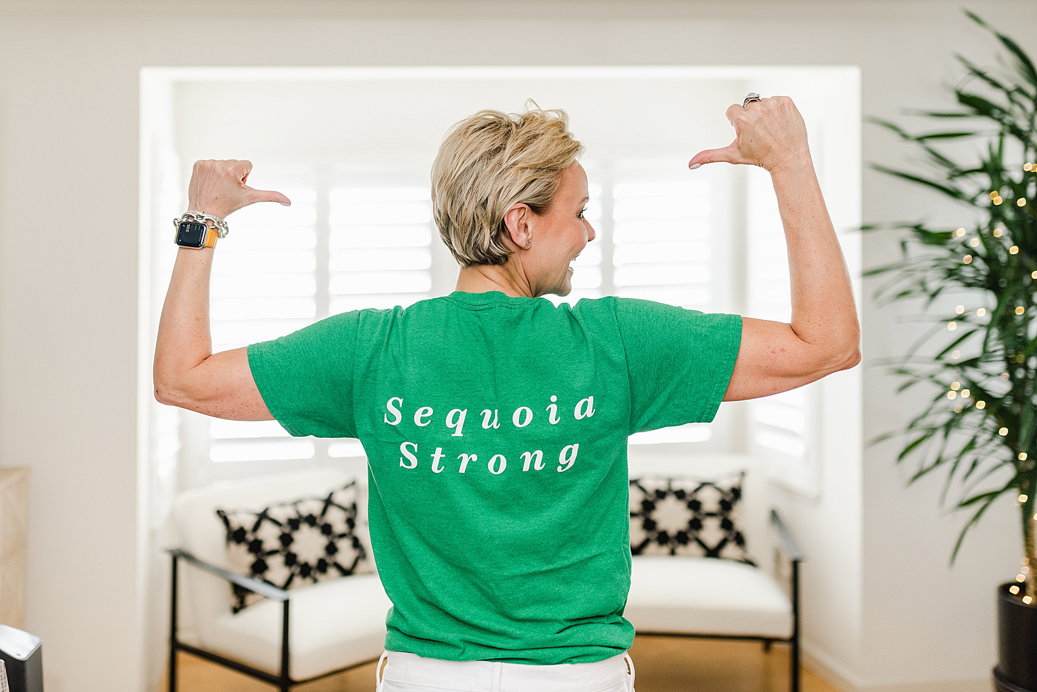 Woman standing with her back to the camera is wearing a green shirt that says "Sequoia Strong" while her arms are raised and her thumbs pointing to the shirt.