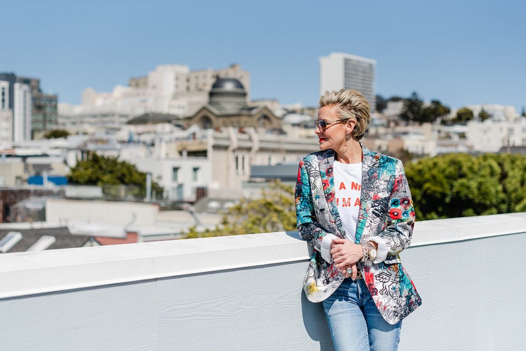 Woman wearing a colorful printed blazer is standing by a balcony ledge overlooking the city. She's leaning on the ledge and is looking at the view.