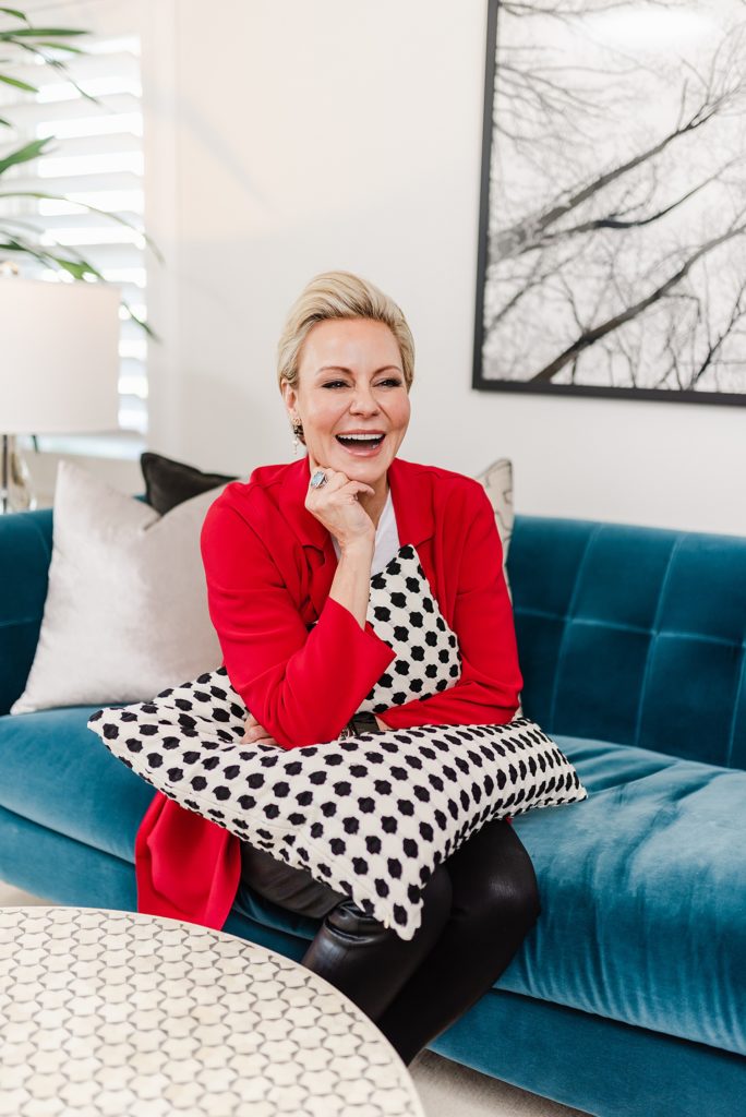 Laughing woman in red blazer is sitting on a blue couch and has a black and white throw pillow on her lap. She is leaning her chin on her hand while her elbow is on the pillow on her lap.