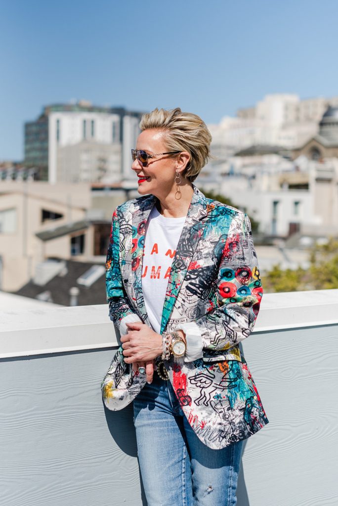 Woman wearing a colorful printed blazer is standing by a balcony ledge overlooking the city. She's leaning on the ledge and is looking at the view.