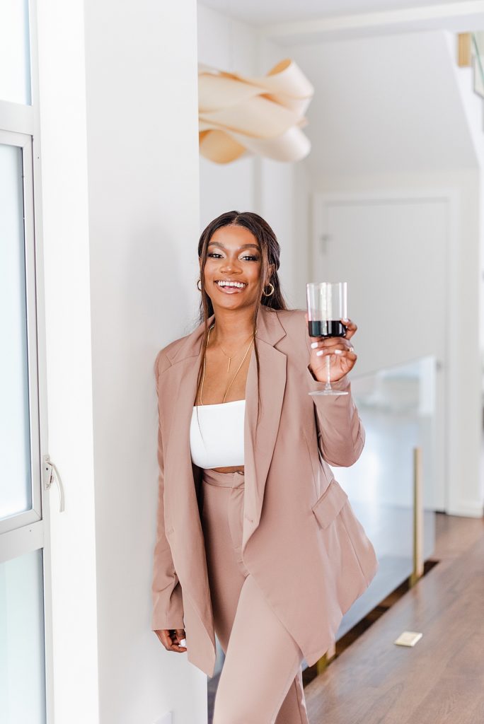woman wearing a beige coat and slacks leaning on the wall while holding a wine glass with one hand. the glass is 1/3 of the way full.