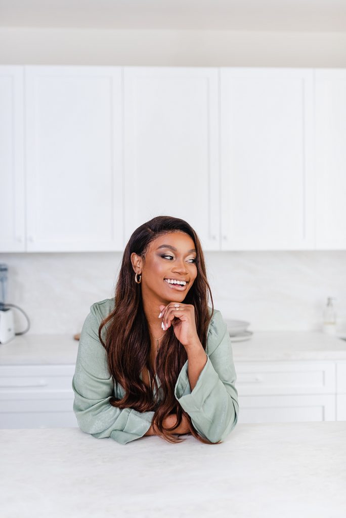 smiling woman leaning on a countertop while looking to the side. her knuckles are lightly pressed under her chin as if lifting her face with her hand.