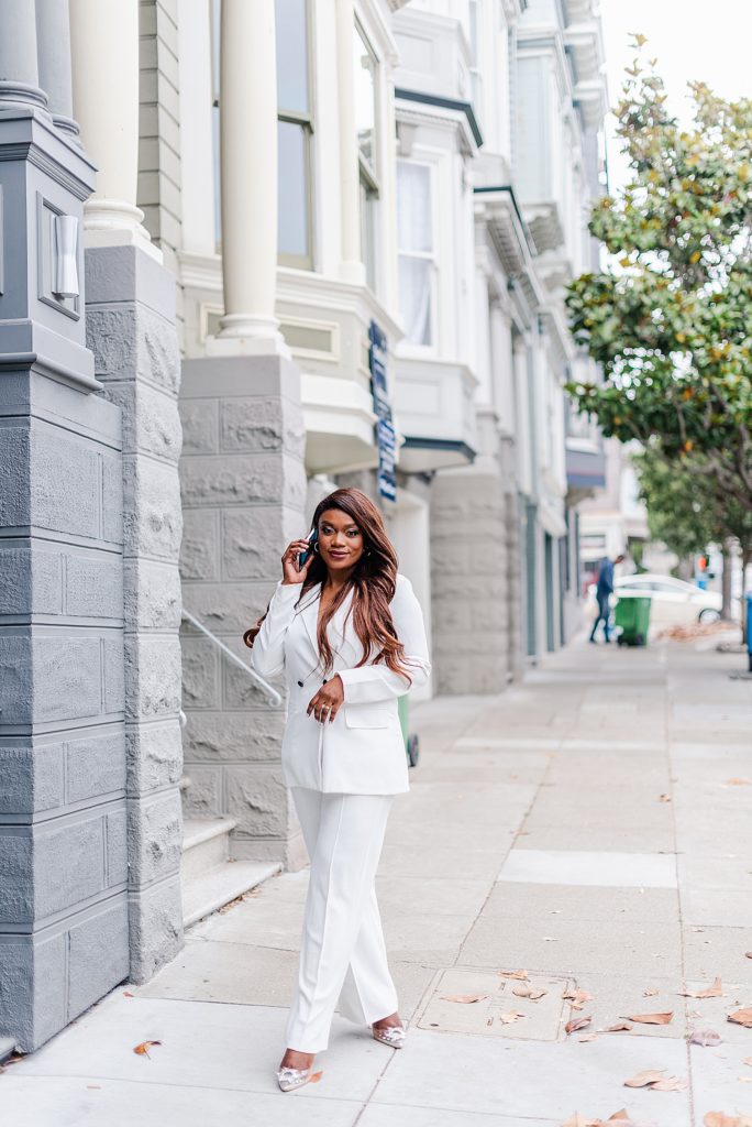 woman in a white suit is standing on the sidewalk while holding a phone to her ear