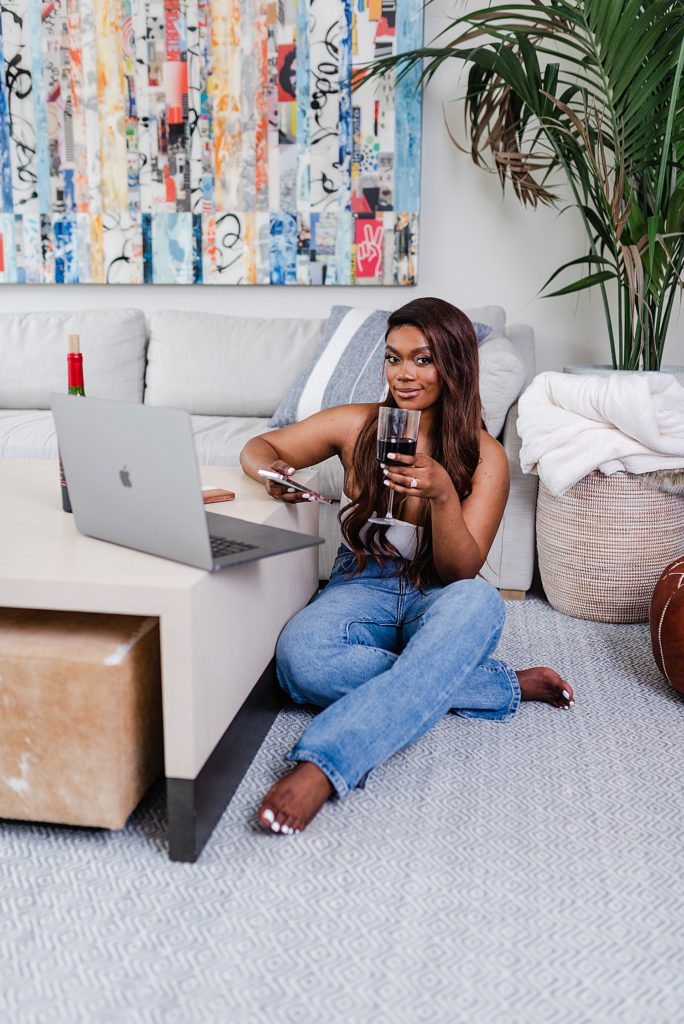 woman is sitting on the floor comfortably while holding a glass of wine on one hand and her phone on the other. she is sitting beside a coffee table with an open laptop facing her on top of it