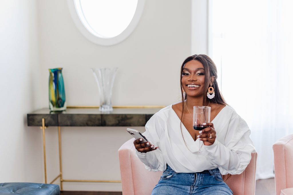 smiling woman sitting on a pink chair while holding a glass of wine and her phone
