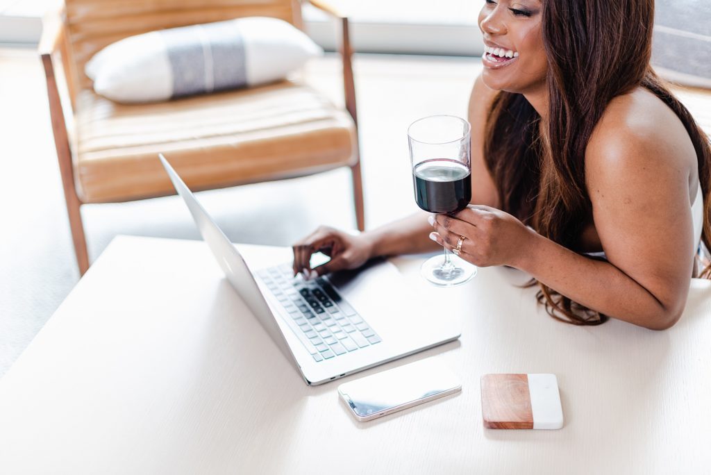 smiling woman holding a glass of wine while facing her laptop
