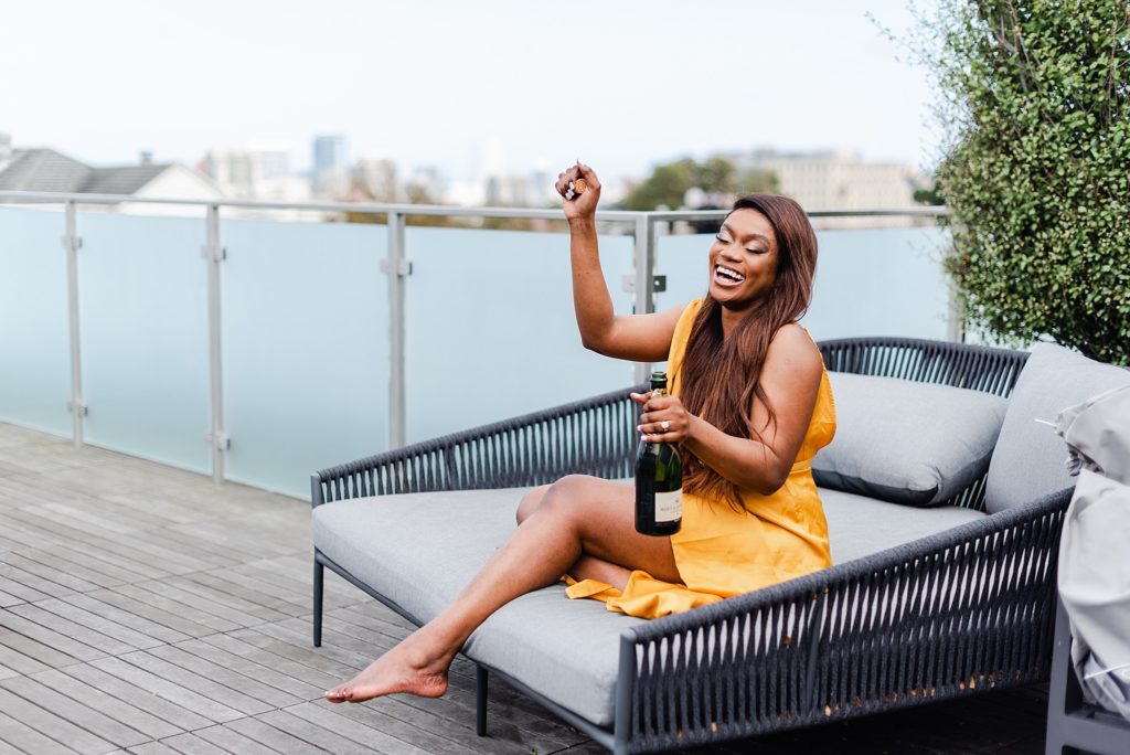 laughing woman is lounging on an outdoor daybed while holding a bottle of wine