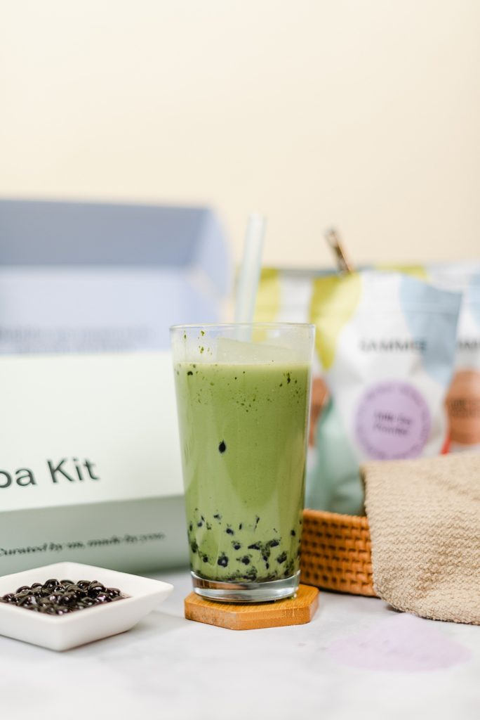 Matcha green milk tea in a clear glass on a coaster, a saucer of boba is beside the glass and SAMMEE products are in the background