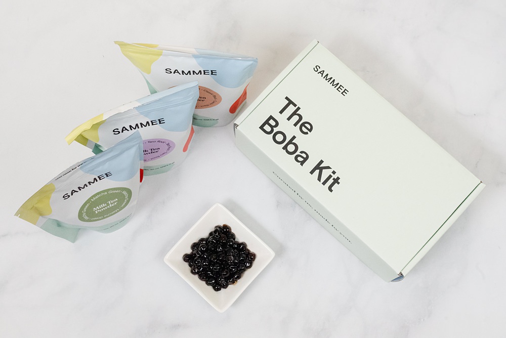 flatlay image of 3 Sammee milk tea powder packs beside The Boba Kit box, and a saucer with cooked boba