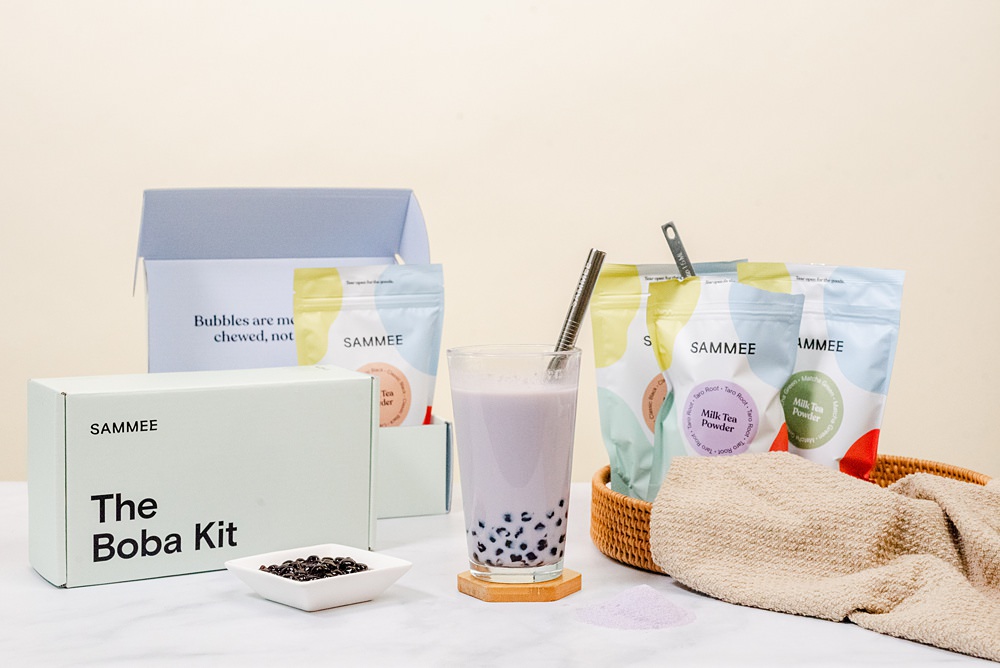 Taro root milk tea in a clear glass on a coaster, The Boba Kit box and a saucer of boba is beside the glass, and SAMMEE products are in the background