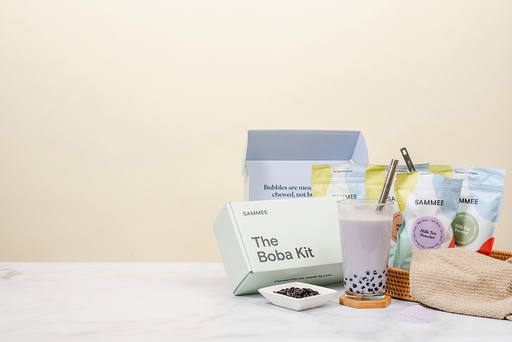 Taro root milk tea in a clear glass on a coaster, The Boba Kit box behind it, a saucer of boba beside the glass in front of the box, and SAMMEE milk tea powders on the other side of the glass. There's also an open box of The Boba Kit in the background